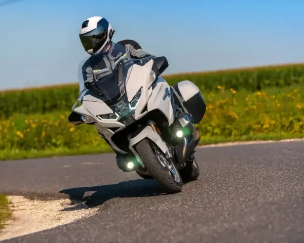 Motocyklista w kasku i kombinezonie pochyla się na zakręcie na białym motocyklu BMW R 1250 RT, przemierzając malowniczą drogę wśród pól.