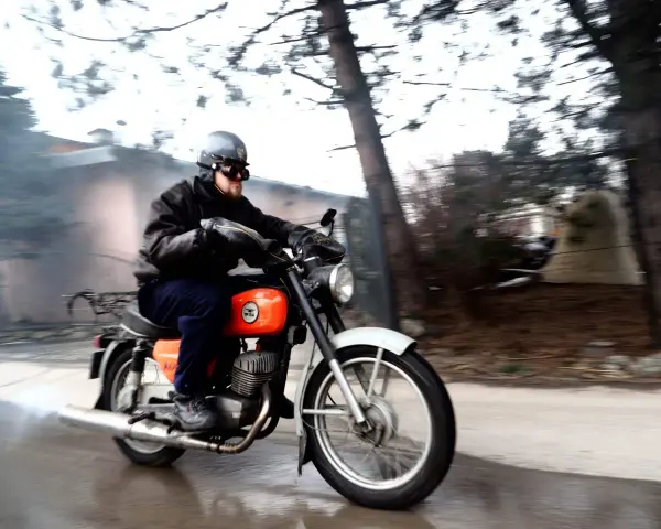 Mężczyzna w kasku i goglach pędzi na pomarańczowym motocyklu WSK 125. Widać dym z rury wydechowej, sugerujący dużą prędkość maksymalną.