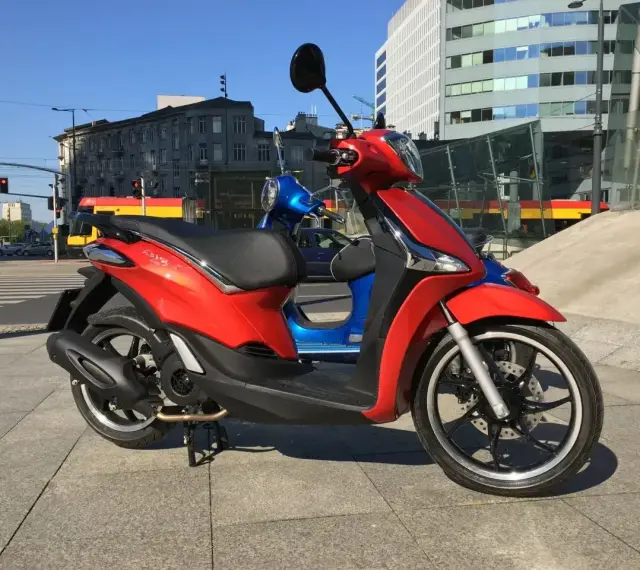 Czerwony Piaggio Liberty 125, obok niebieskiego, na tle miasta. Idealny do sprawdzenia piaggio liberty 125 dane techniczne.