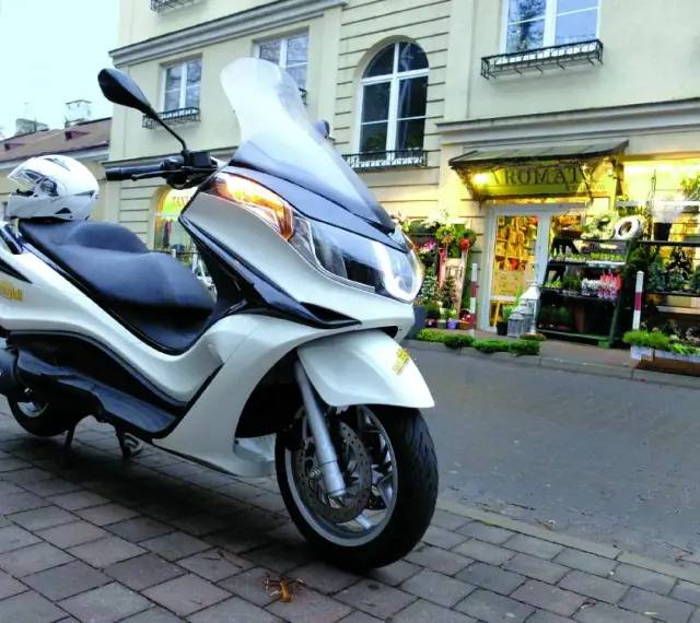 Biały skuter Piaggio X10 125, z kaskiem na siedzeniu, zaparkowany przed kwiaciarnią. Dane techniczne tego modelu są imponujące.