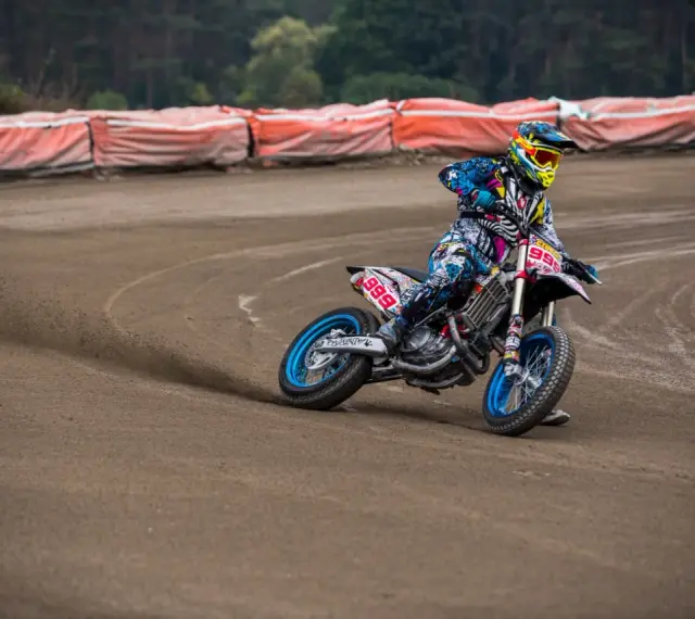 Motocyklista w kolorowym stroju śmiga po piaszczystym torze **flat track**, pochylony w zakręcie.
