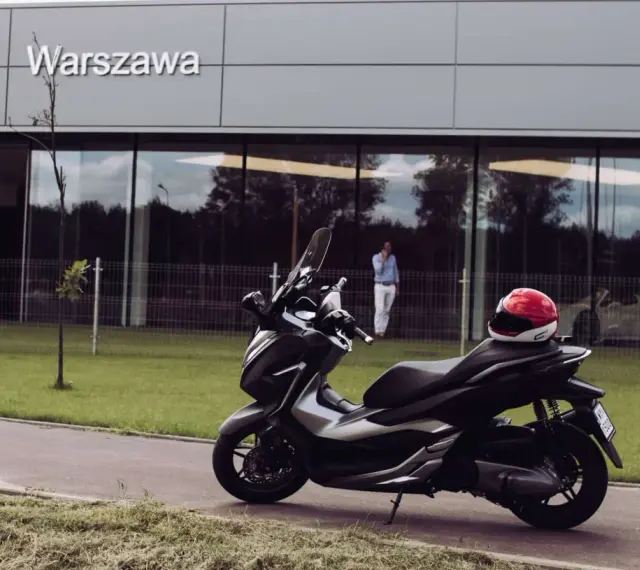 Elegancki skuter Honda Forza 300 zaparkowany przed budynkiem z napisem "Warszawa". Czerwony kask leży na siedzeniu.