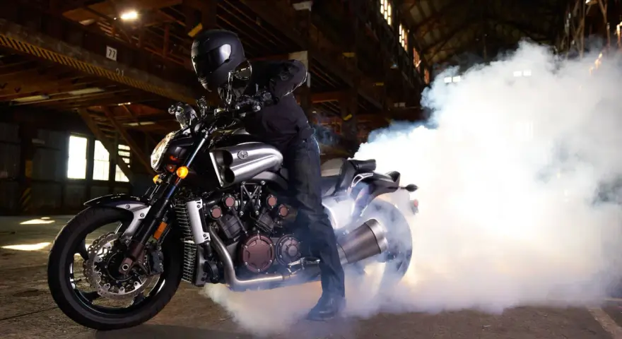 Motocyklista w kasku na Yamasze V-Max wykonuje burnout, tworząc kłęby dymu w opuszczonej hali.