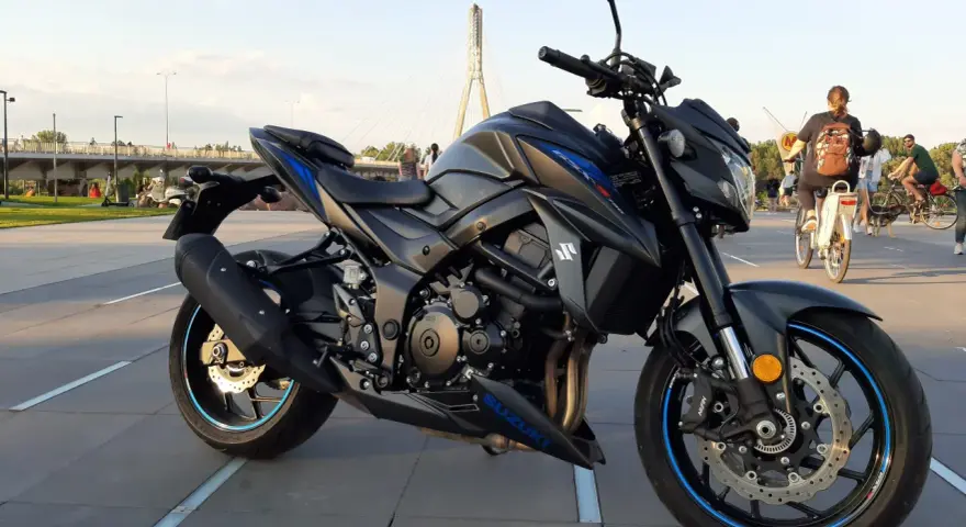 Czarny motocykl Suzuki GSX-S 750 z niebieskimi akcentami na felgach i owiewkach, zaparkowany na placu z widocznym w tle mostem i ludźmi.