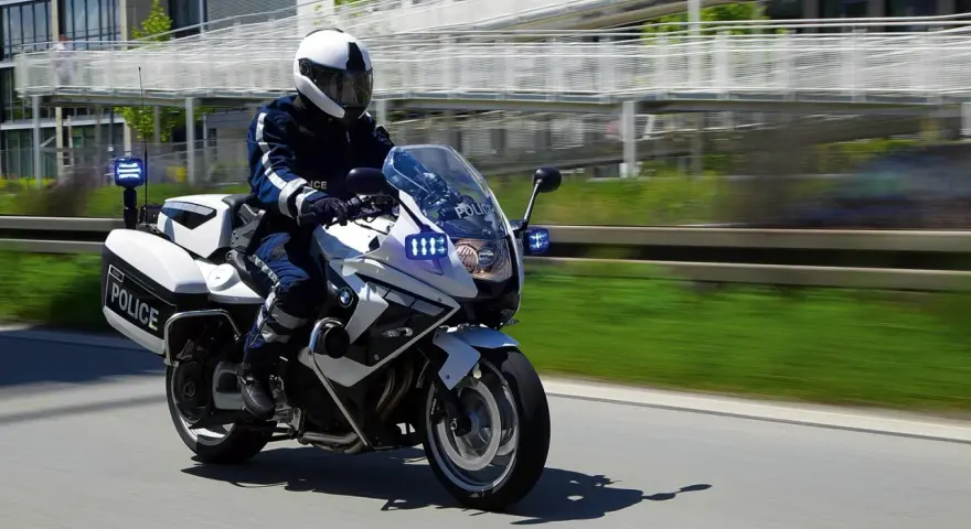 Policyjny motocykl BMW F800GT z włączonymi sygnałami świetlnymi pędzi po drodze.