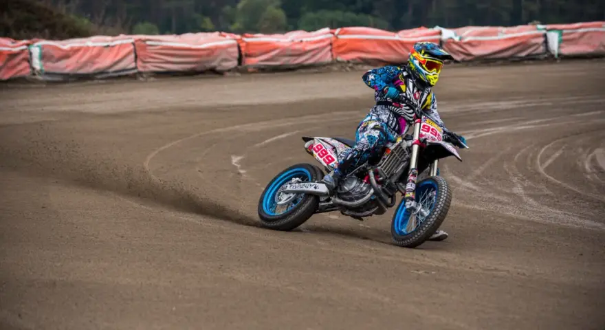 Motocyklista w kolorowym stroju śmiga po piaszczystym torze **flat track**, pochylony w zakręcie.