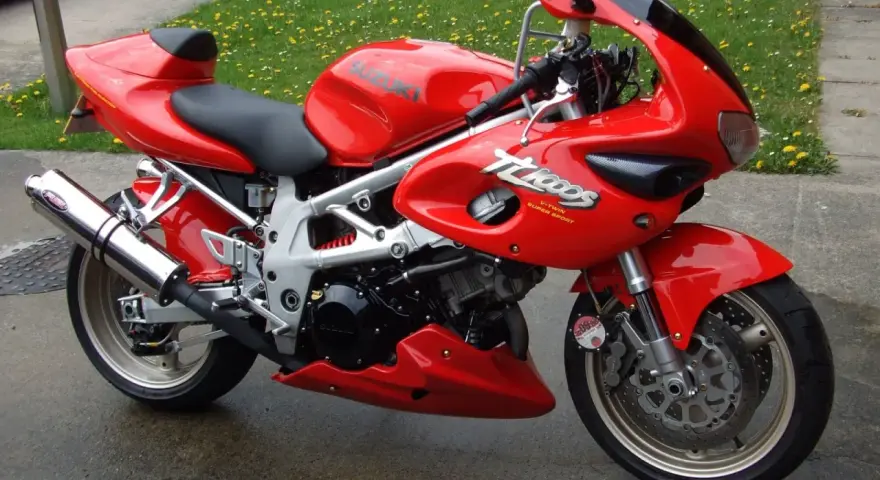 Czerwony motocykl Suzuki TL1000 z napisem "V-TWIN SUPER SPORT" na owiewce, zaparkowany na mokrej nawierzchni obok trawnika.