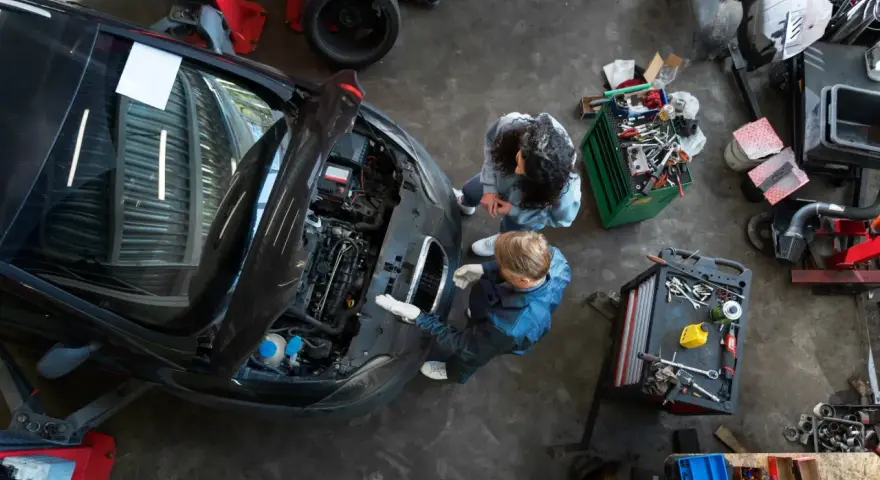 Warsztat samochodowy z dwoma mechanikami przy otwartej masce czarnego auta i narzędziami.