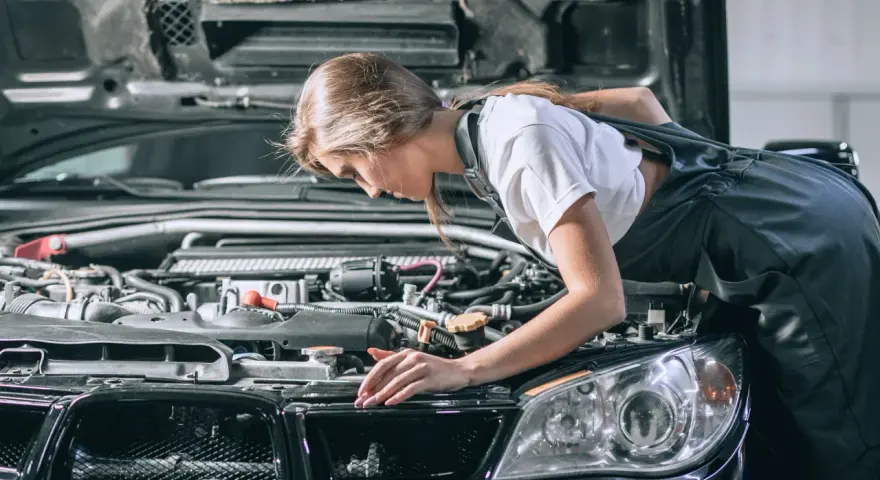 Młoda mechaniczka pochyla się nad silnikiem, sprawdzając go. To może być początek jej drogi, jak otworzyć warsztat samochodowy.