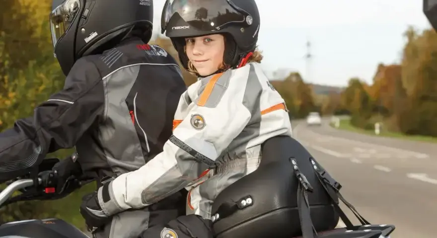 Uśmiechnięte dziecko na motocyklu, siedzące przed dorosłym, w kasku i kurtce motocyklowej.