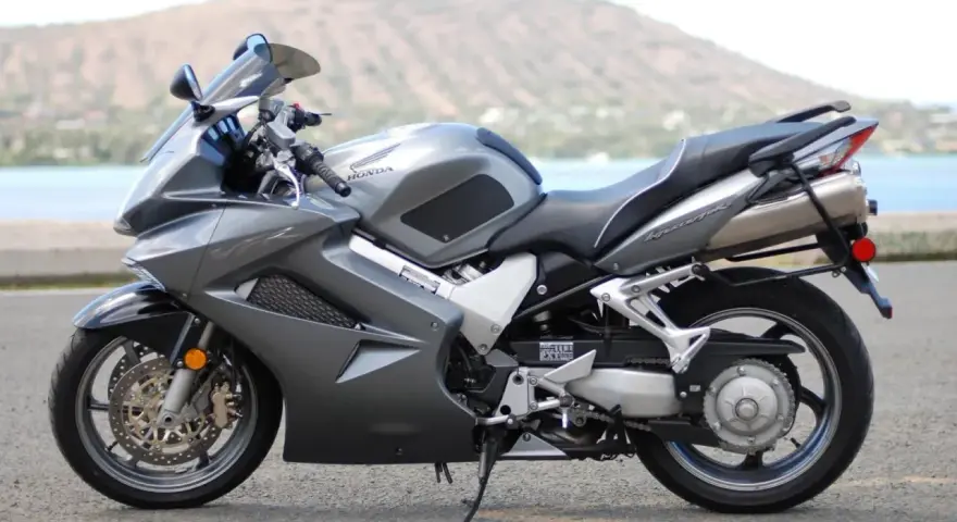 Szary motocykl Honda VFR 800 z widokiem na wodę i wzgórza w tle.