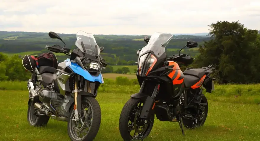 Dwa motocykle turystyczne, w tym pomarańczowy KTM 1290 Super Adventure, stoją na trawie z widokiem na zielone wzgórza.
