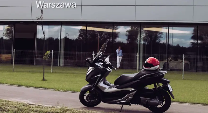 Elegancki skuter Honda Forza 300 zaparkowany przed budynkiem z napisem "Warszawa". Czerwony kask leży na siedzeniu.