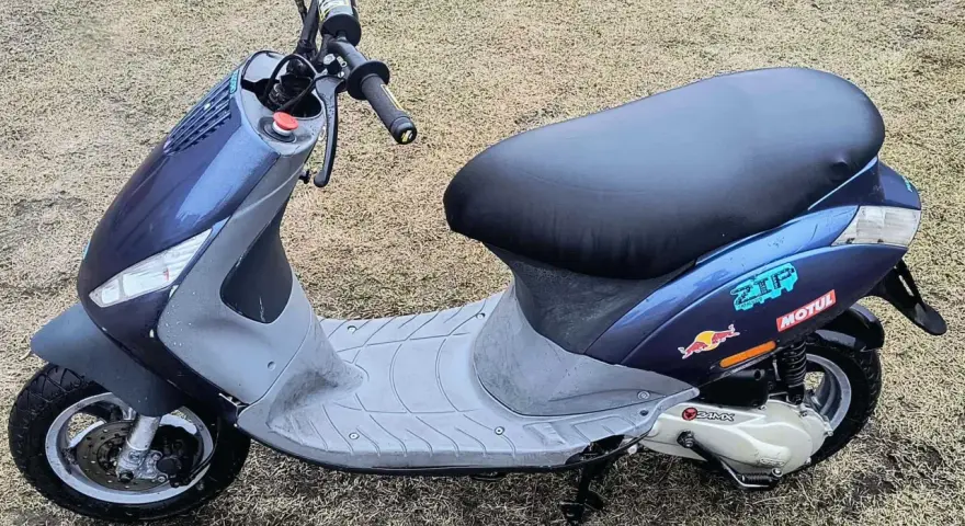 Niebieski skuter z naklejkami Red Bull i Motul na tle trawy.