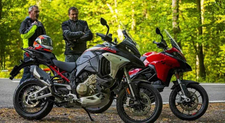 Dwóch motocyklistów z kaskiem na siedzeniu obok dwóch motocykli Ducati Multistrada, szary i czerwony, na tle leśnej drogi.