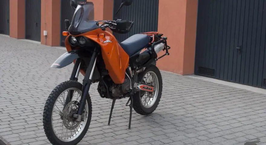 Pomarańczowy motocykl KTM 640 Adventure stoi na brukowanym podjeździe przed garażami.