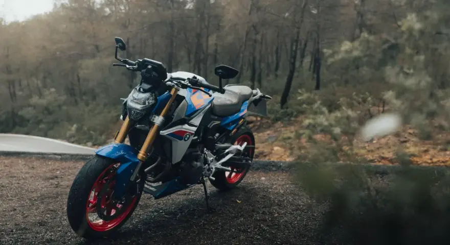 Niebiesko-biały motocykl BMW F900R z czerwonymi felgami na tle jesiennego lasu.