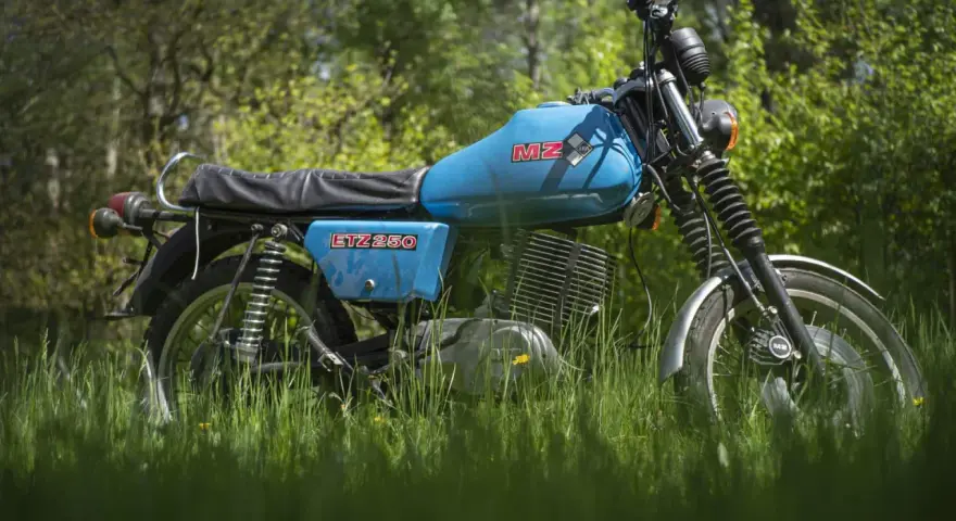 Niebieski motocykl MZ ETZ 250 stojący na trawie, z drzewami w tle.