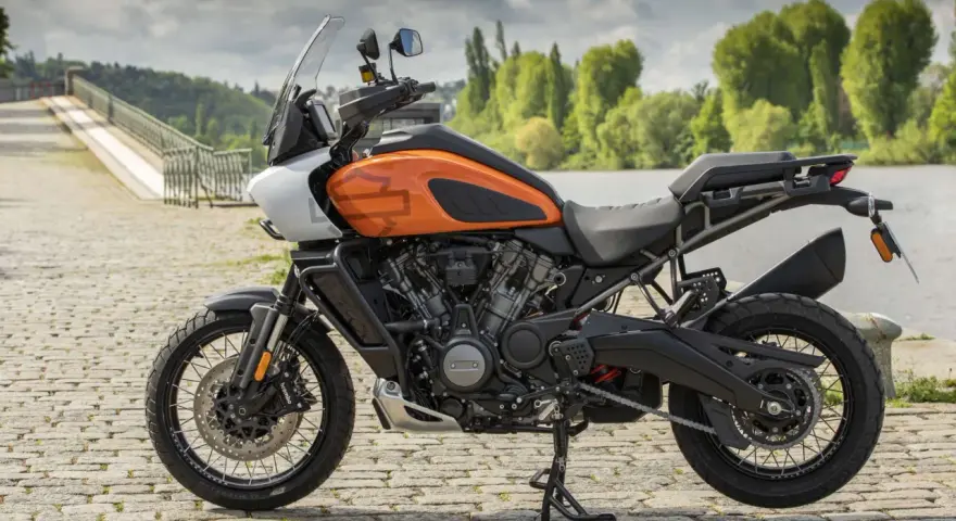 Pomarańczowy Harley-Davidson Pan America gotowy na przygodę, z mostem i zielenią w tle.