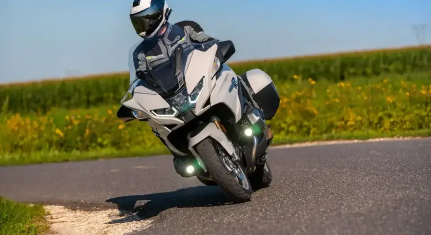 Motocyklista w kasku i kombinezonie pochyla się na zakręcie na białym motocyklu BMW R 1250 RT, przemierzając malowniczą drogę wśród pól.