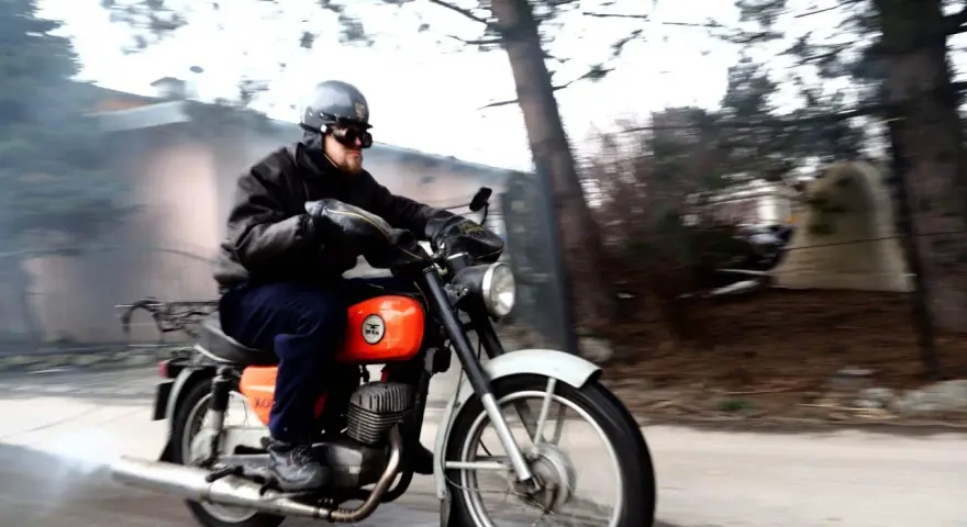 Mężczyzna w kasku i goglach pędzi na pomarańczowym motocyklu WSK 125. Widać dym z rury wydechowej, sugerujący dużą prędkość maksymalną.