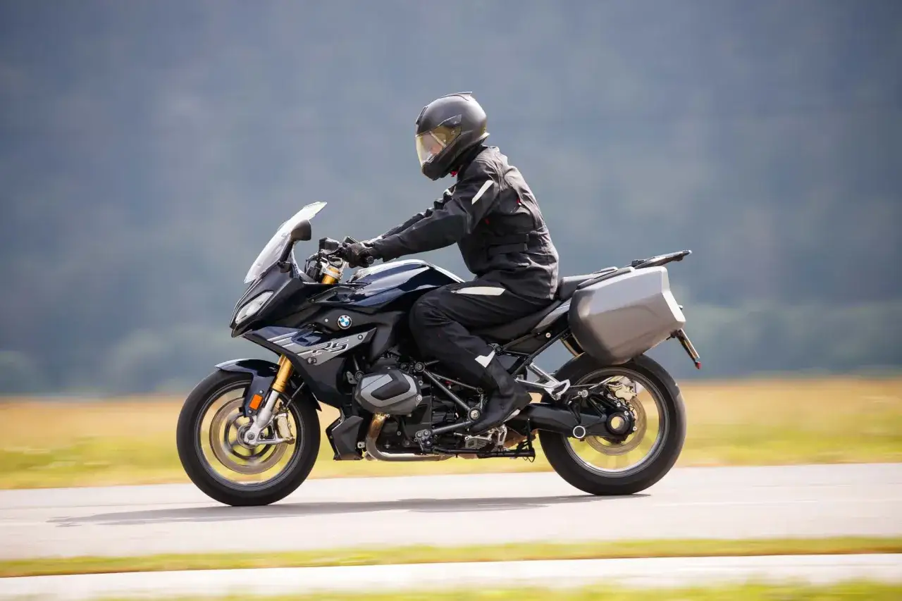 Motocyklista w kasku i kombinezonie pędzi na czarnym BMW 1250 RS po drodze, z bagażnikami po bokach.
