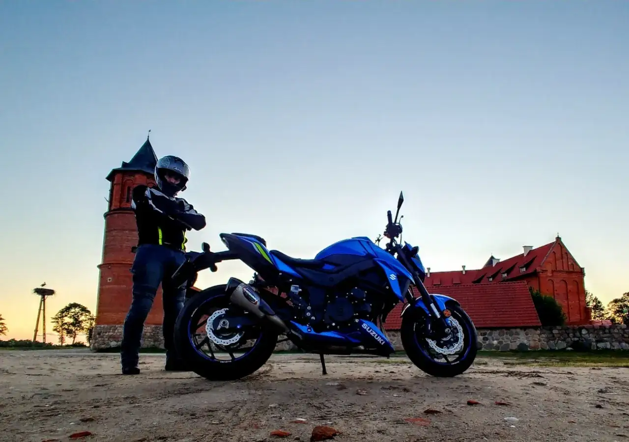 Motocyklista w kasku obok niebieskiego Suzuki GSX-S 750. W tle zabytkowa wieża i czerwony budynek. Suzuki GSX-S 750 dane techniczne.