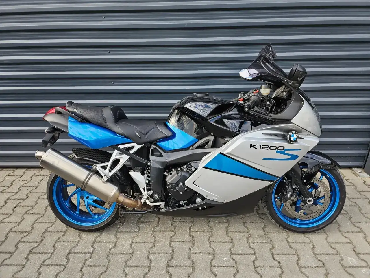 Sportowy motocykl BMW K1300GT w kolorze czarno-srebrno-niebieskim, zaparkowany na chodniku przed szarą, falistą ścianą.