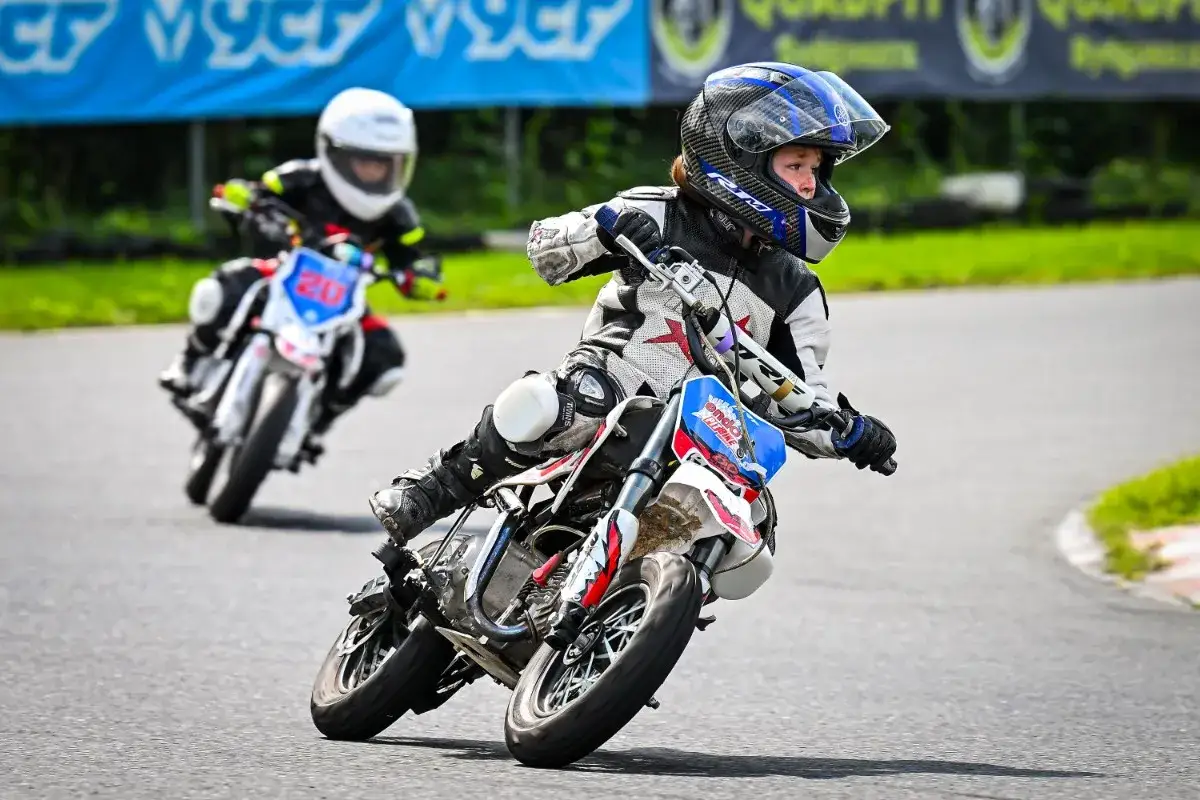 Dzieci ścigają się na motocyklach na torze wyścigowym. Młodzi zawodnicy w kaskach i kombinezonach zmagają się na zakręcie, pokazując swoje umiejętności w wyścigach flat track.
