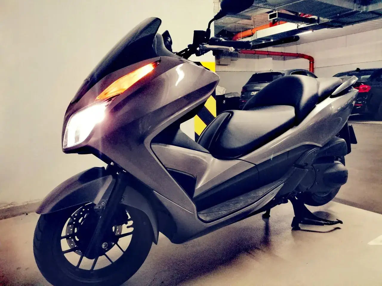 Szary skuter Honda Forza 300 z włączonymi światłami, zaparkowany w garażu podziemnym.