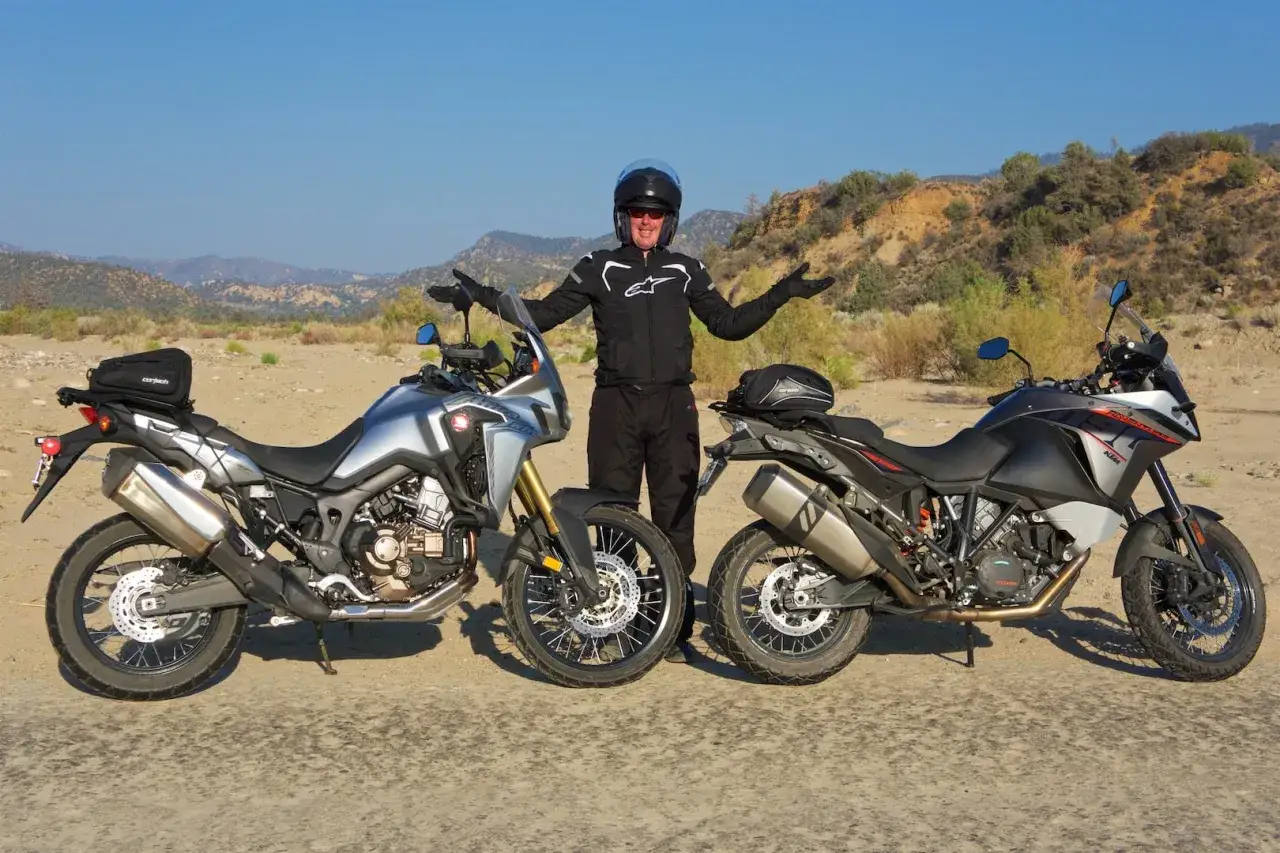 Motocyklista w kasku prezentuje dwa motocykle: srebrną Hondę Africa i czarno-szary motocykl KTM.