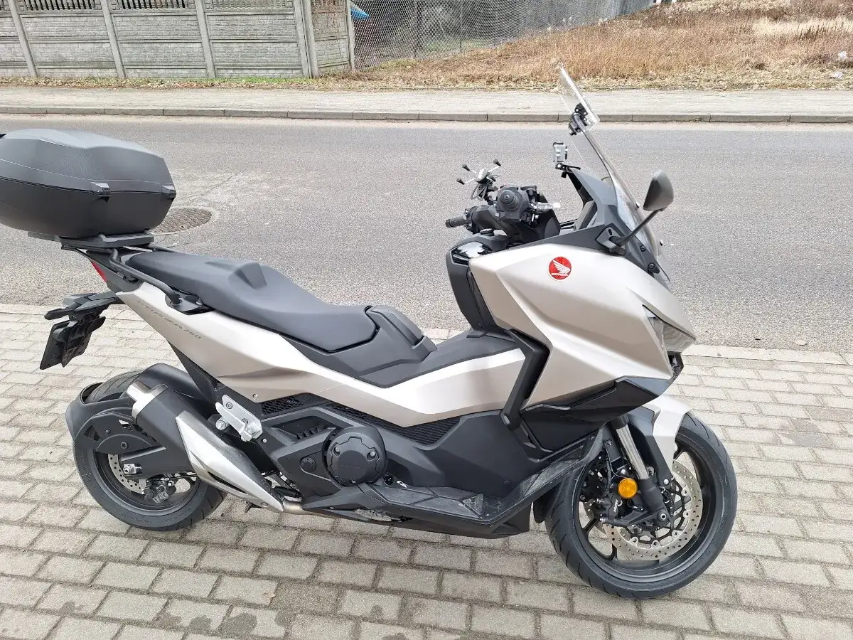 Złoty skuter Honda Forza 750, gotowy do osiągnięcia swojej maksymalnej prędkości. Z kuferkiem na bagaż i sportowym wyglądem.