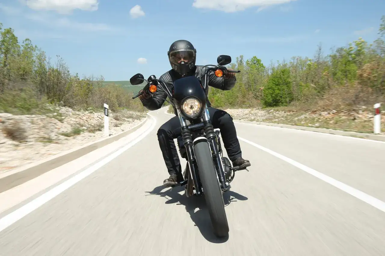Motocyklista w kasku i skórzanej kurtce pędzi na motocyklu Harley Davidson Iron 1200 po krętej drodze wśród zieleni.