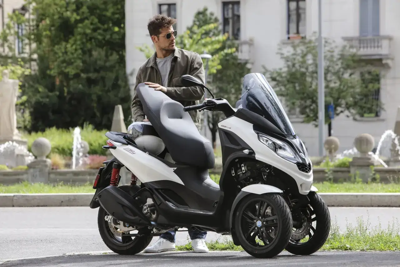 Mężczyzna w okularach opiera się o biały Piaggio MP3 300. Poznaj jego piaggio mp3 300 dane techniczne i poczuj wolność.