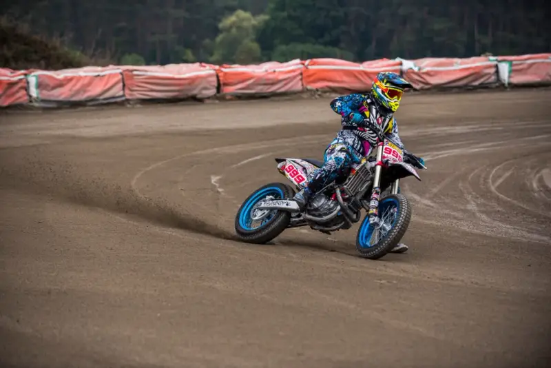 Motocyklista w kolorowym stroju śmiga po piaszczystym torze **flat track**, pochylony w zakręcie.