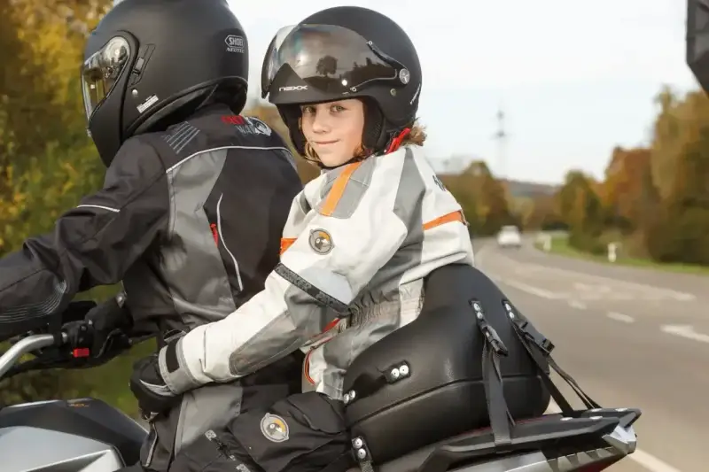 Uśmiechnięte dziecko na motocyklu, siedzące przed dorosłym, w kasku i kurtce motocyklowej.